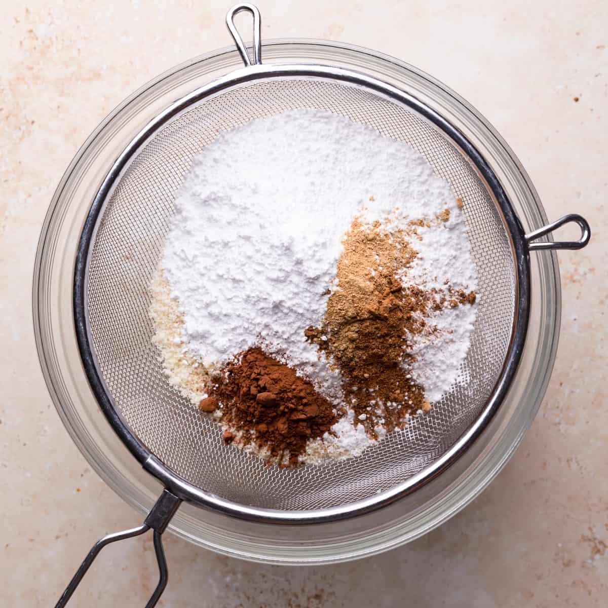 a baker gently sifting almond flour and cocoa powder into a bowl, hands visible