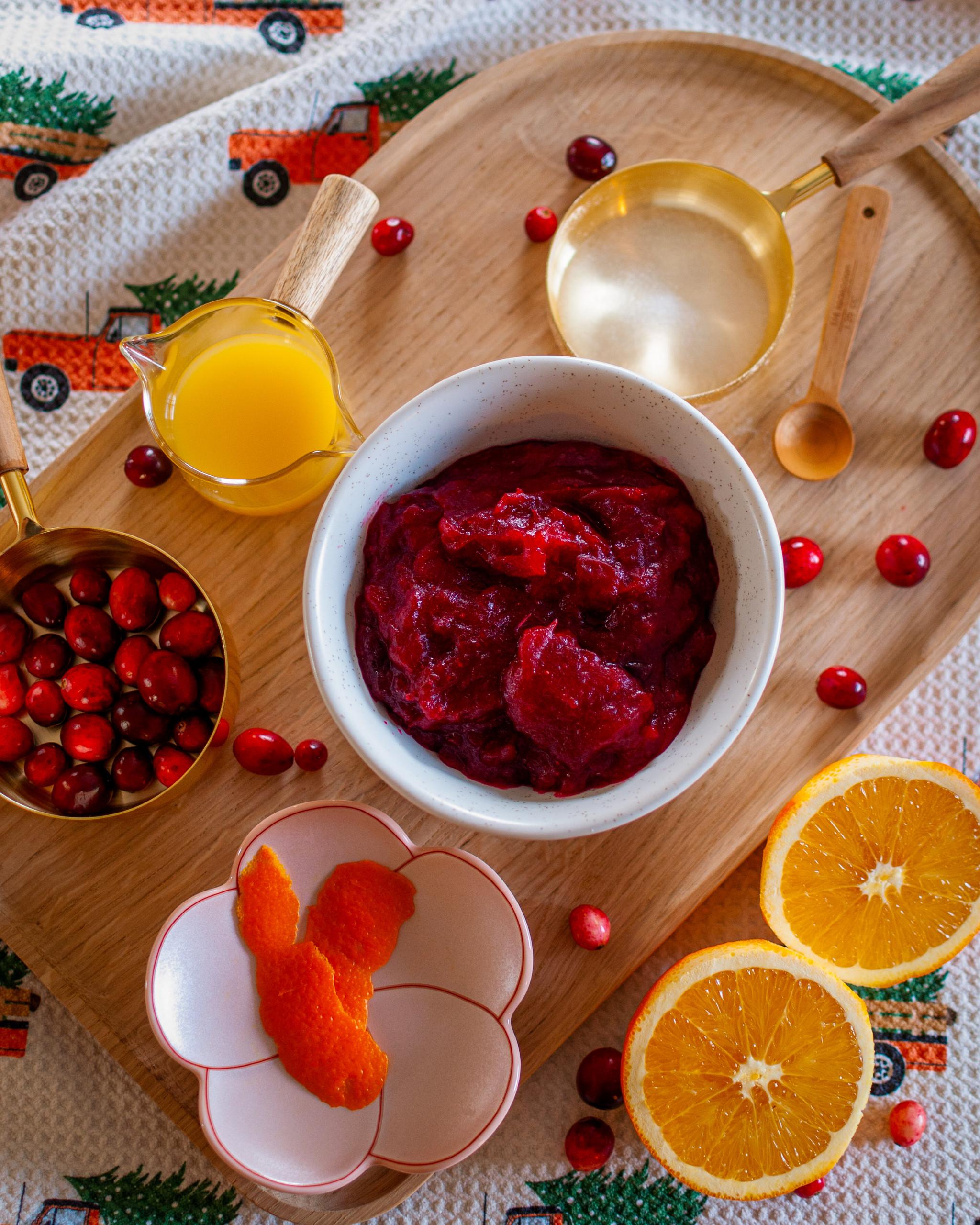 A vibrant bowl of homemade cranberry sauce, garnished with fresh cranberries and an orange peel, on a rustic wooden table with holiday decorations.