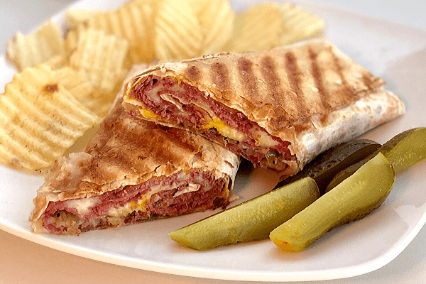 a hand holding a pastrami and swiss wrap, with a bite taken out, showcasing the melted cheese and pastrami
