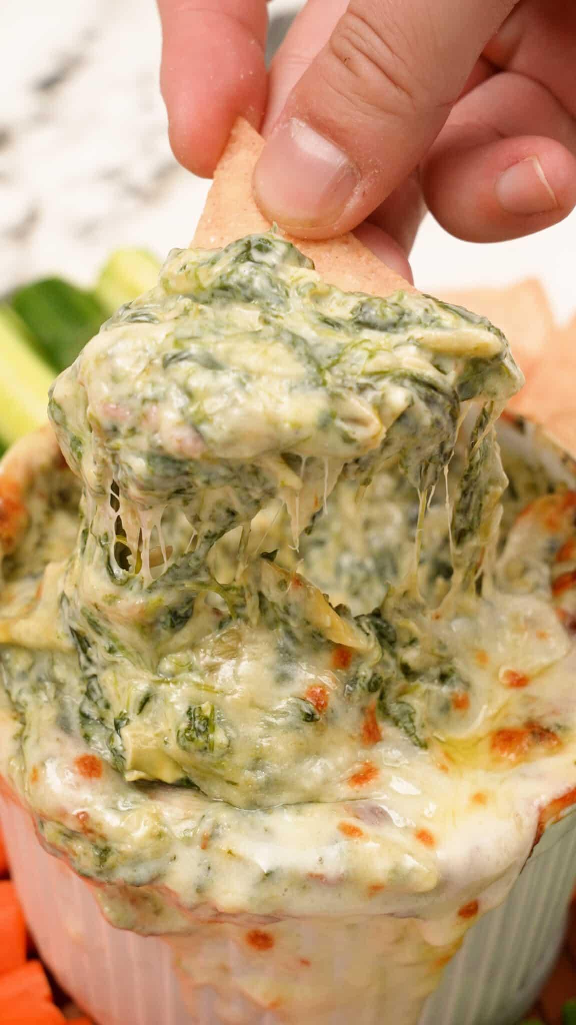 Close-up of creamy spinach artichoke dip being scooped with a cracker, showing melted cheese pull