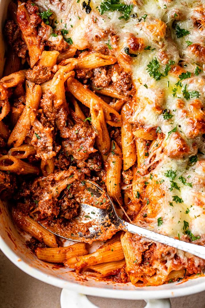 Ingredients for baked ziti laid out on a kitchen counter: pasta, sauce, cheeses, ground meat, and herbs