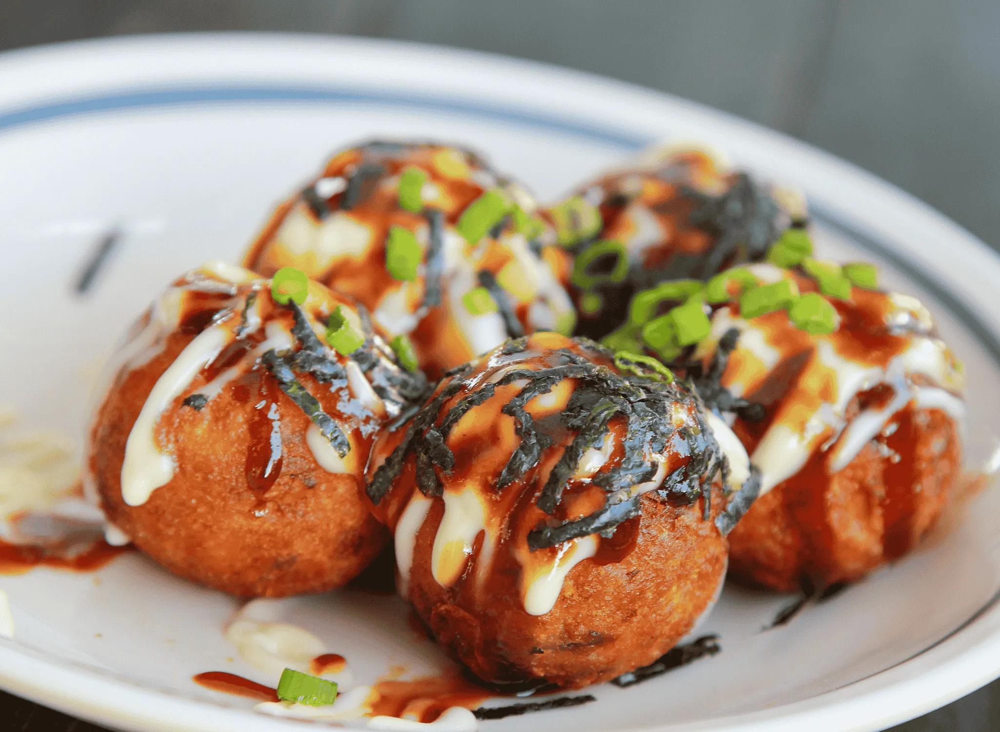 A close-up shot of several Yuzu Kosho Yawp Takoyaki balls arranged on a plate, drizzled with sauce and garnished with green onions and nori seaweed flakes