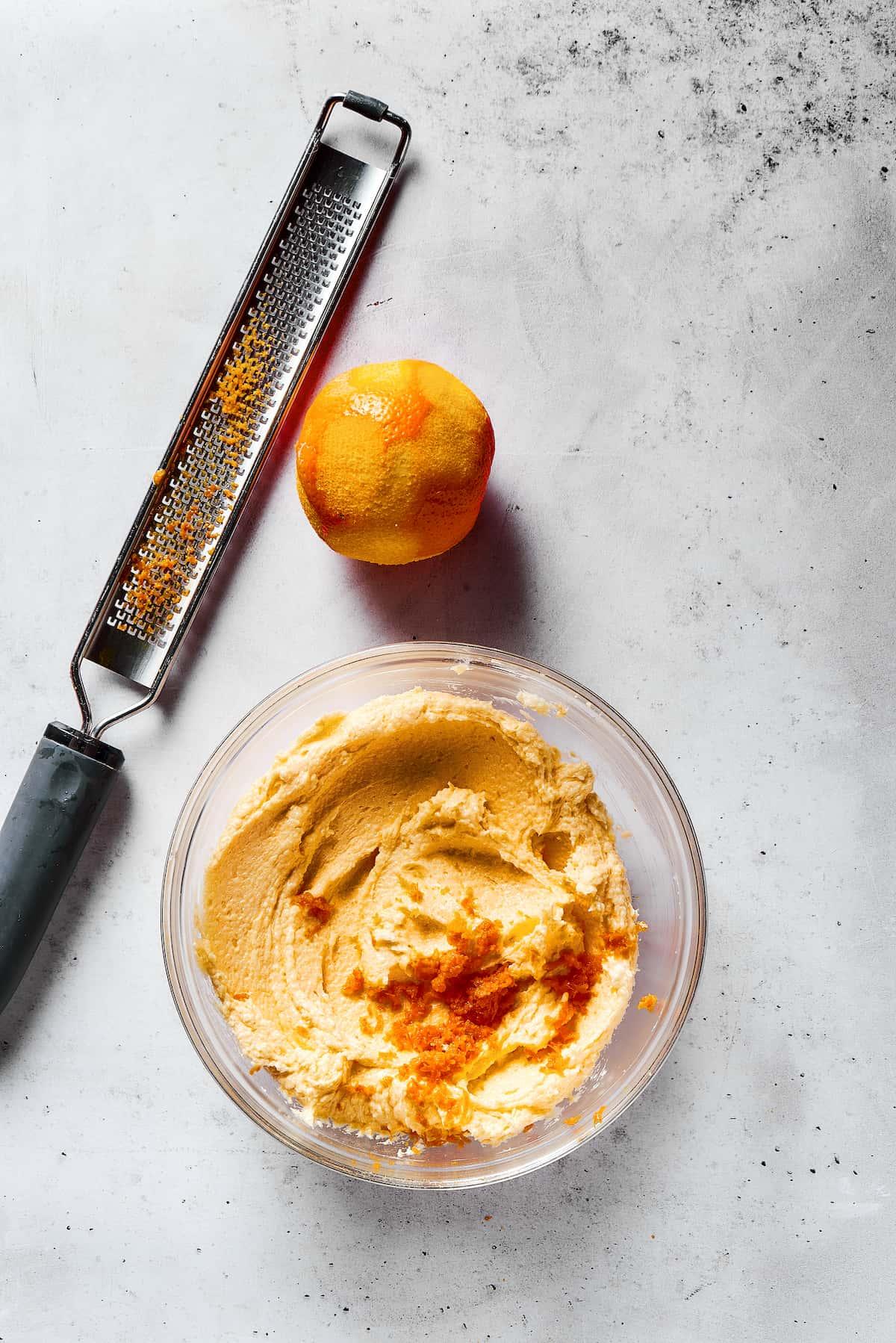 Close-up of a hand zesting a fresh orange over a bowl of cookie dough