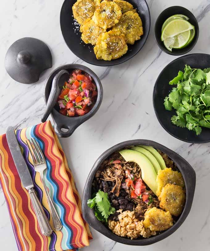 A plate of plantain burritos surrounded by colorful ingredients like avocados, tomatoes, and cilantro