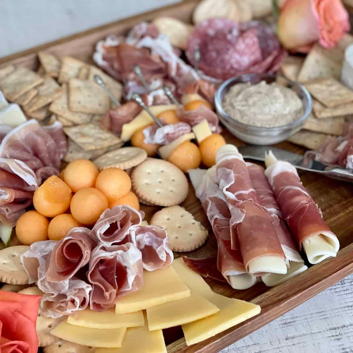 Charcuterie board with prosciutto roses and fig jam in a ramekin, a hand reaching for a piece of cheese
