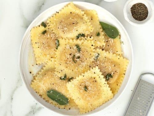 Close-up shot of a perfectly plated butternut squash ravioli dish with visible steam, showcasing the fresh sage and butter sauce