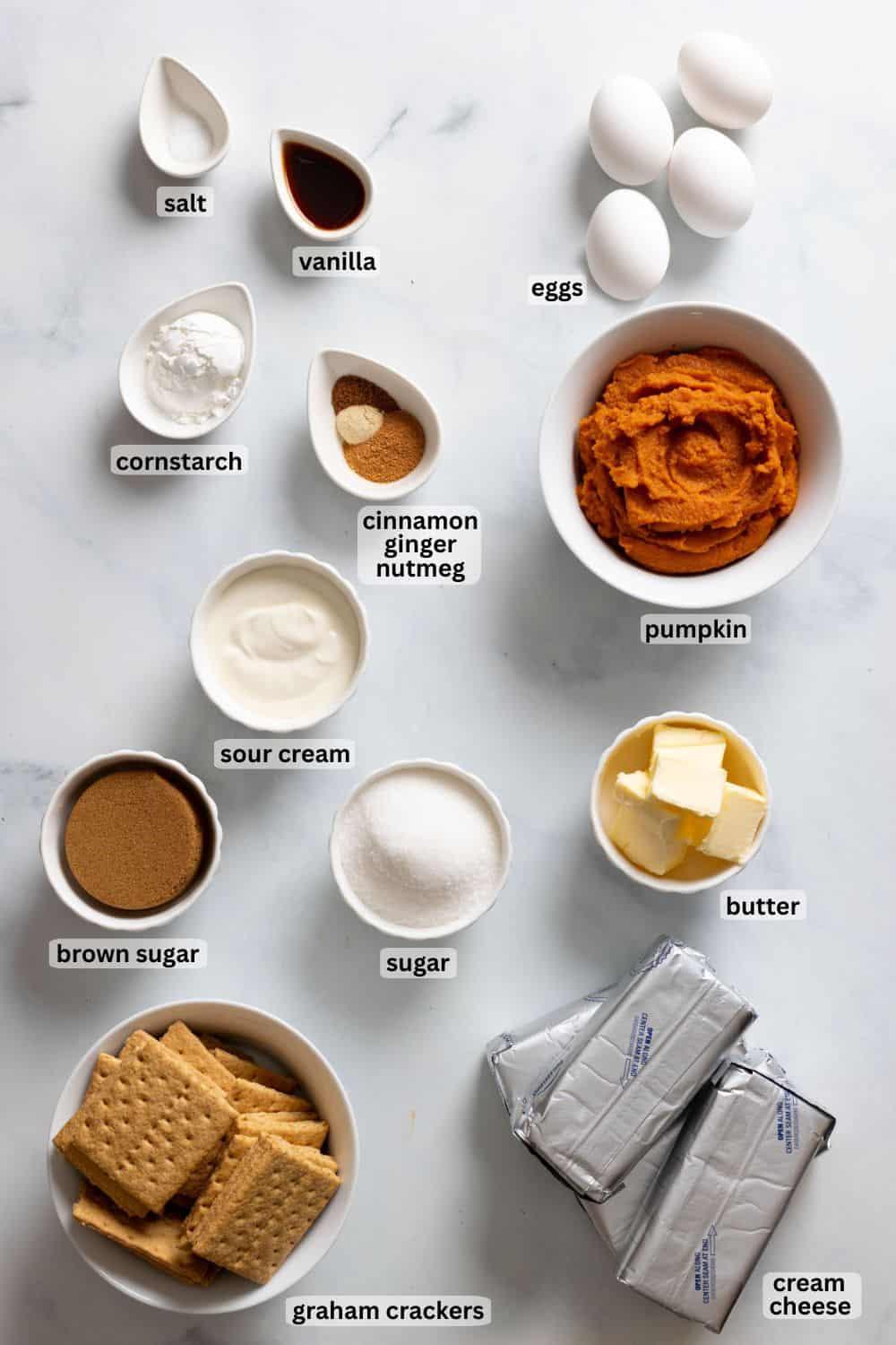 Ingredients for pumpkin cheesecake laid out on a wooden table, including pumpkin puree, cream cheese, spices, and graham crackers