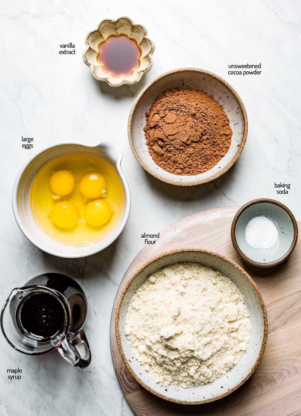 ingredients for almond flour chocolate cake laid out on a kitchen counter: almond flour, cocoa powder, eggs, sugar, oil, vanilla, hot water