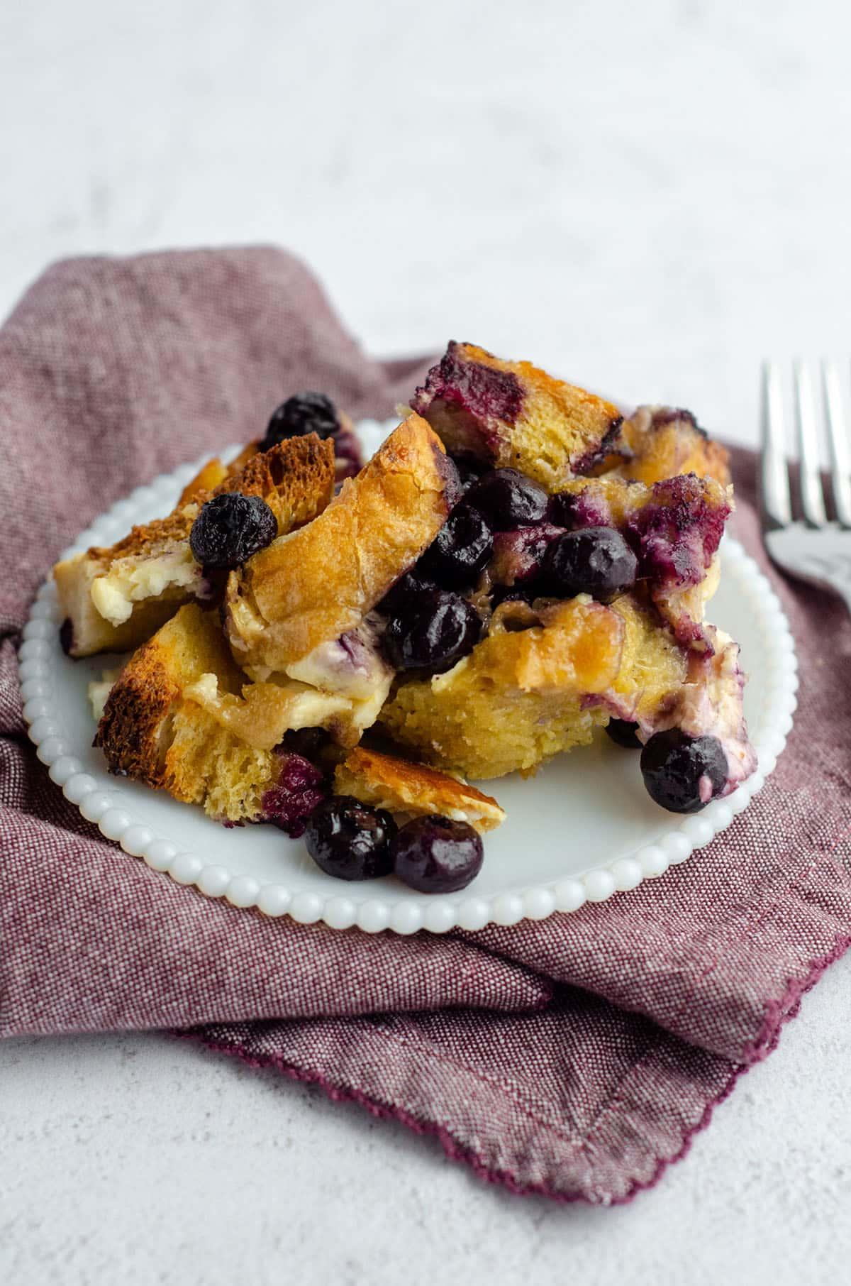 Close-up of a golden brown French toast casserole slice with blueberries spilling out, on a white plate with a fork