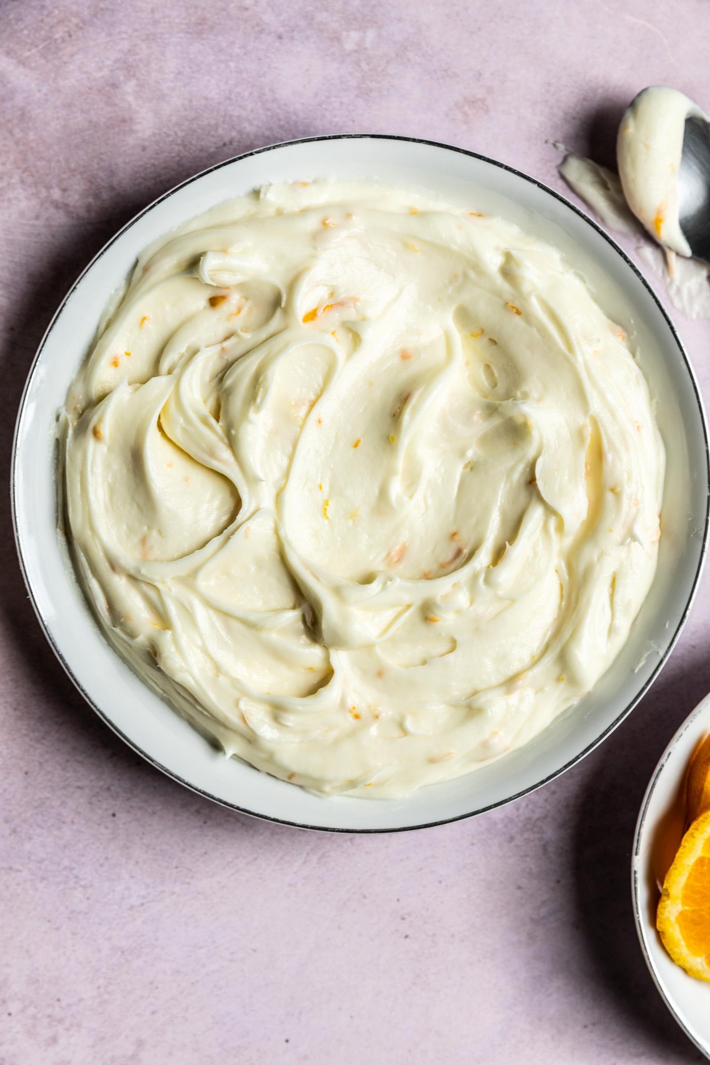bowl of creamy white cream cheese frosting with orange zest being stirred, ready to be spread