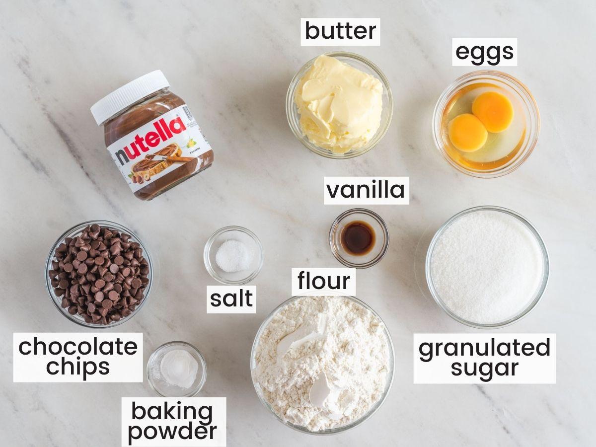 Ingredients for Nutella-stuffed cookies laid out on a kitchen counter: flour, butter, sugar, eggs, Nutella jar