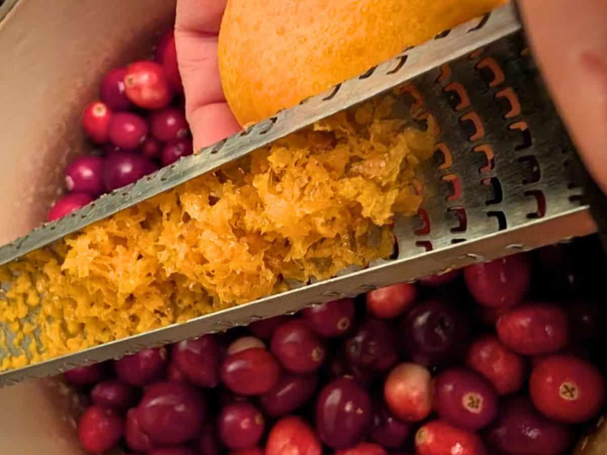 close-up shot of hands zesting a fresh orange over a bowl of sugar, with cranberries nearby