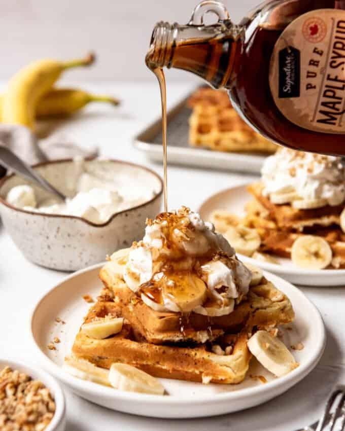 A stack of golden banana pecan waffles dripping with maple syrup, garnished with fresh bananas and chopped pecans