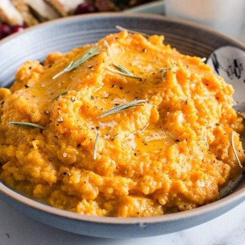 Close-up of creamy mashed sweet potatoes in a bowl, ready to be transferred to a casserole dish, with a hand holding a spatula.