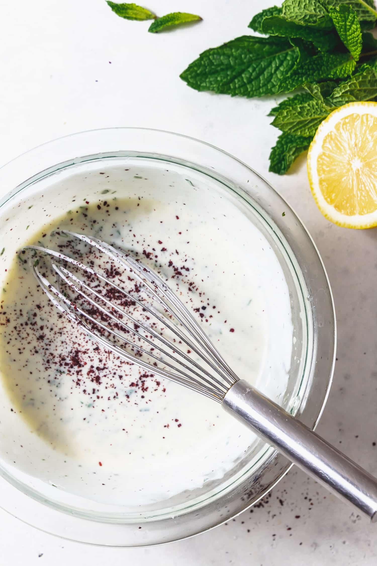 A bowl of creamy sumac yogurt sauce garnished with fresh mint and a sprinkle of sumac