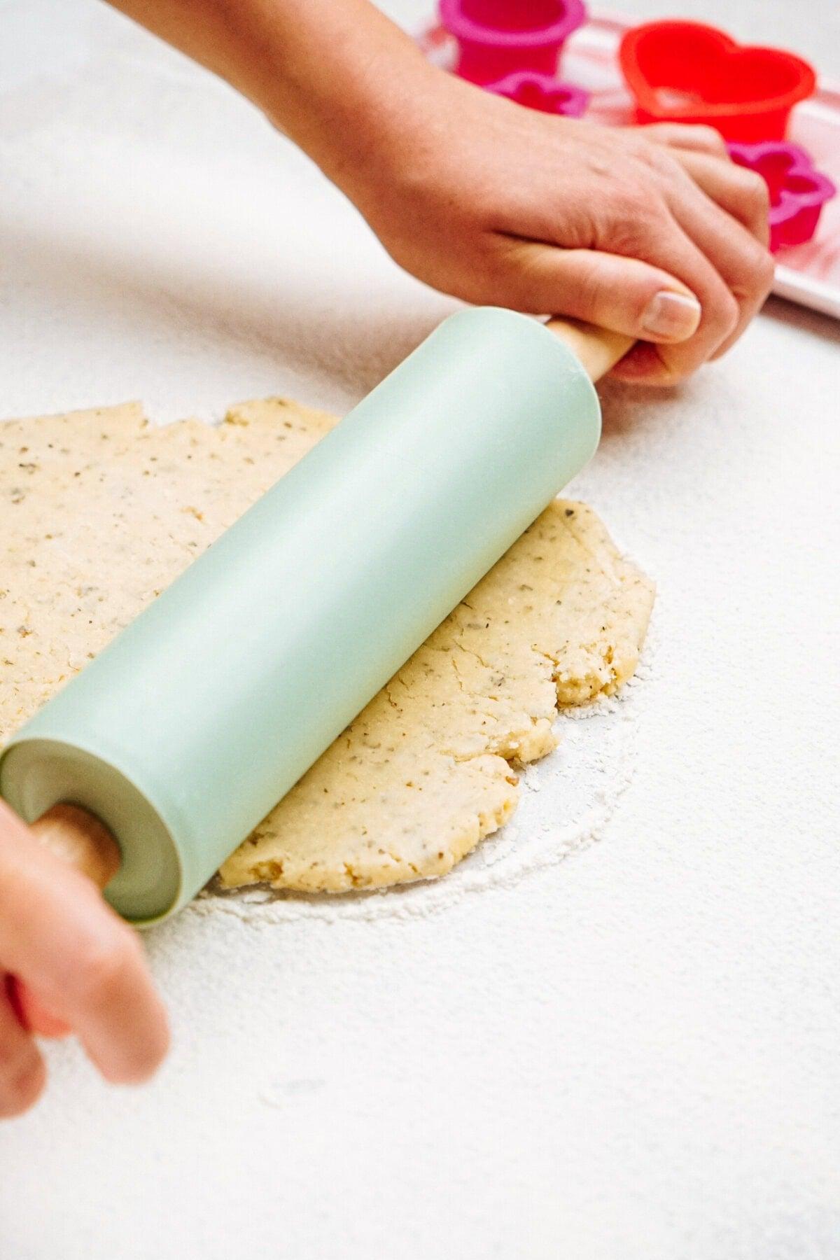 Hands rolling out cheese cracker dough on a lightly floured surface with a rolling pin