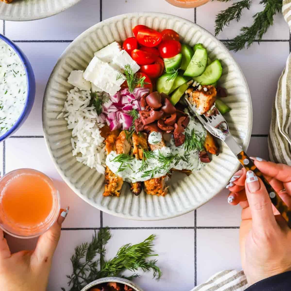 Overhead shot of a beautifully plated Greek chicken bowl on a rustic wooden table with a glass of lemonade in the background