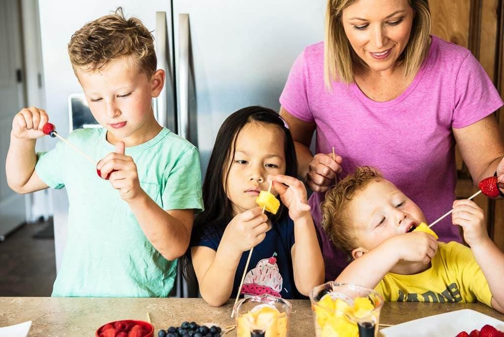a group of kids happily eating Elf Antics Fruit Kabobs, with smiles on their faces and colorful fruit skewers in their hands