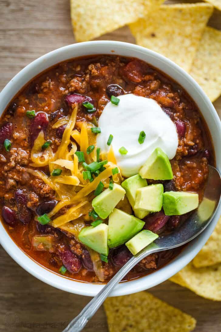 A person garnishing a bowl of slow cooker chili with various toppings