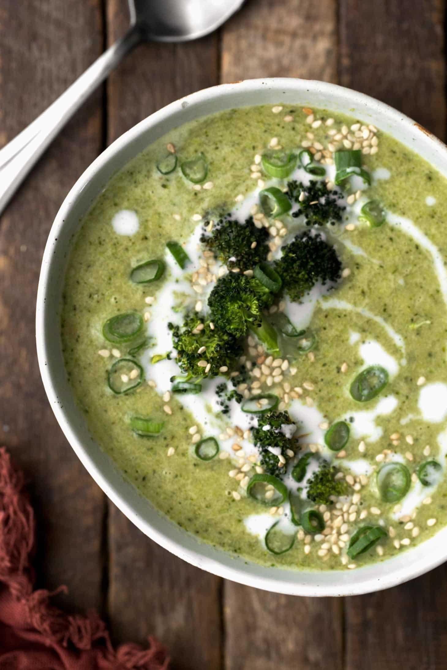 a hand stirring a pot of broccoli and ginger soup with coconut cream being added