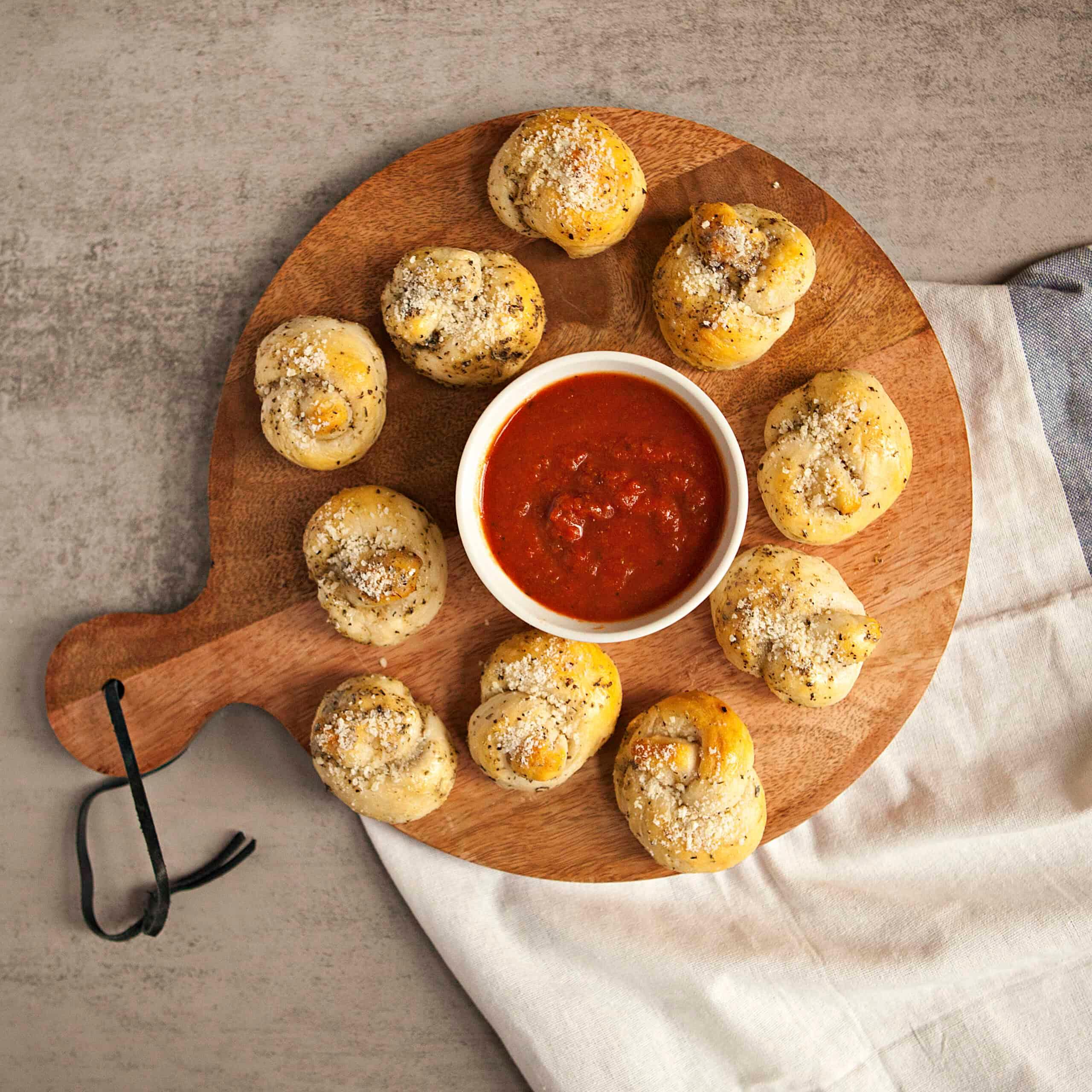 Overhead shot of a rustic wooden board filled with golden cheesy garlic knots, with a bowl of marinara dipping sauce