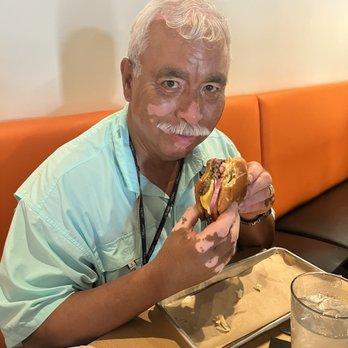 a smiling person holding up a perfectly assembled noodle burger, with a bite taken out to show the juicy filling