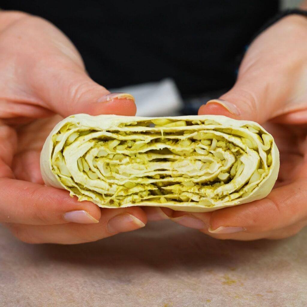 Hands gently rolling up a sheet of puff pastry covered in pesto