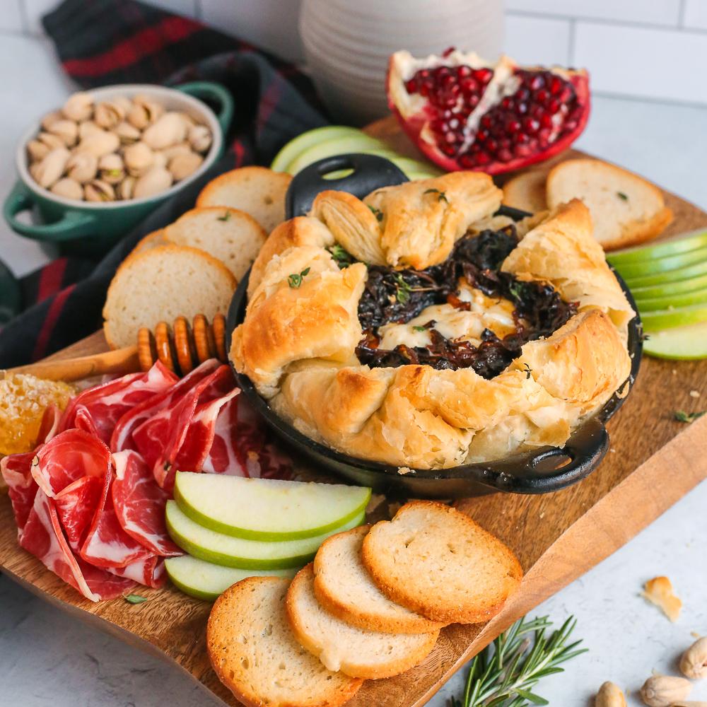 Close-up of a wheel of brie cheese, a block of puff pastry, and some fruit on a wooden cutting board