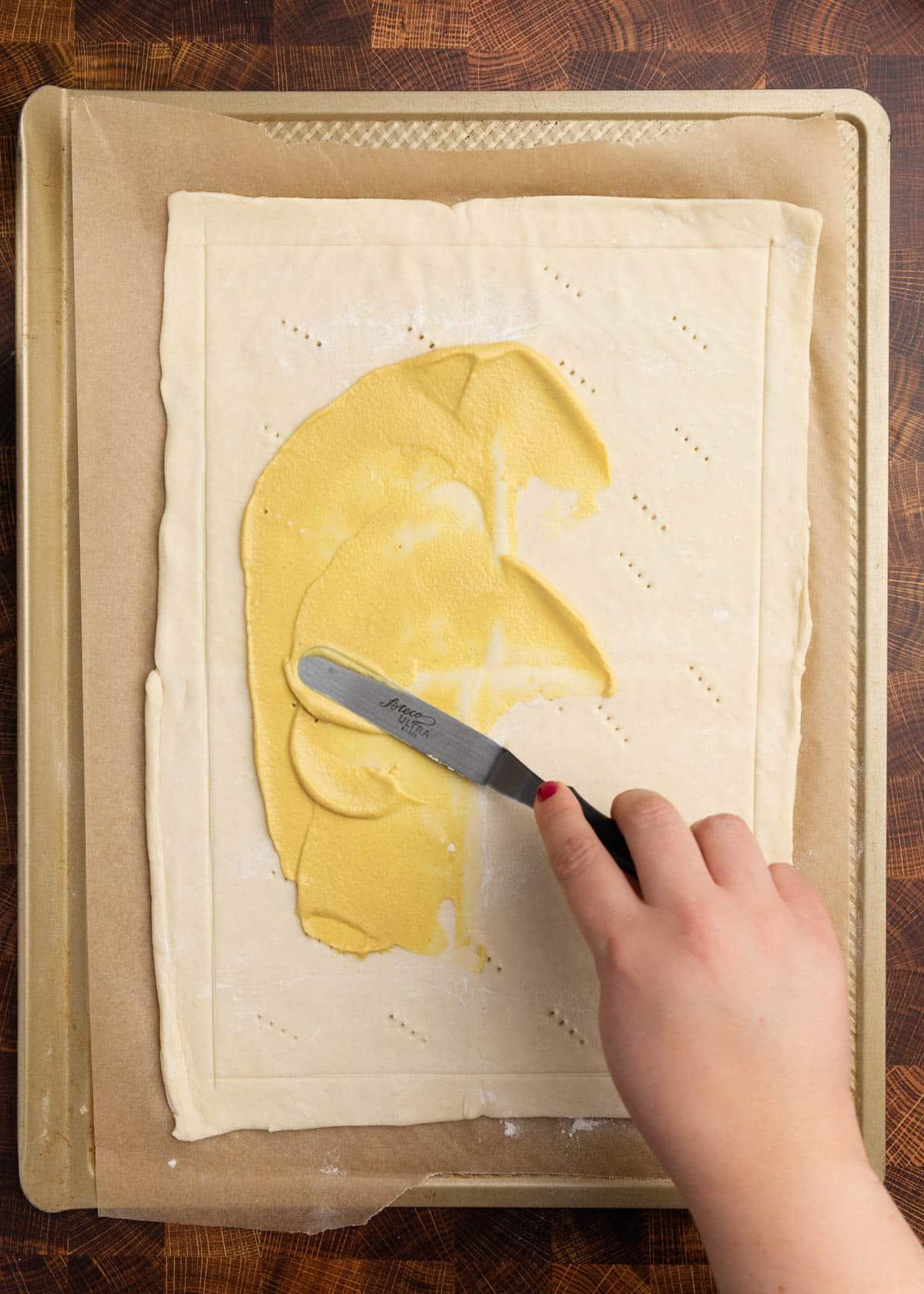 Hands spreading egg wash on puff pastry sheet on parchment paper
