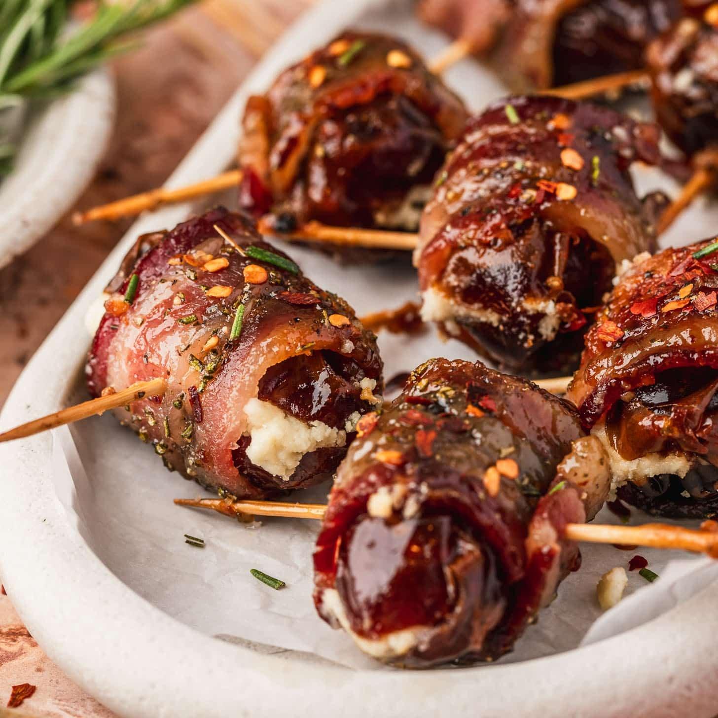 Ingredients laid out for bacon wrapped dates: vibrant Medjool dates, strips of premium thin-cut bacon, a bowl of rich maple syrup, and crumbled goat cheese on a rustic wooden cutting board with a knife nearby.