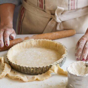Close-up shot of hands gently pressing homemade pastry dough into a tart pan, with flour dust and fresh apples in the background