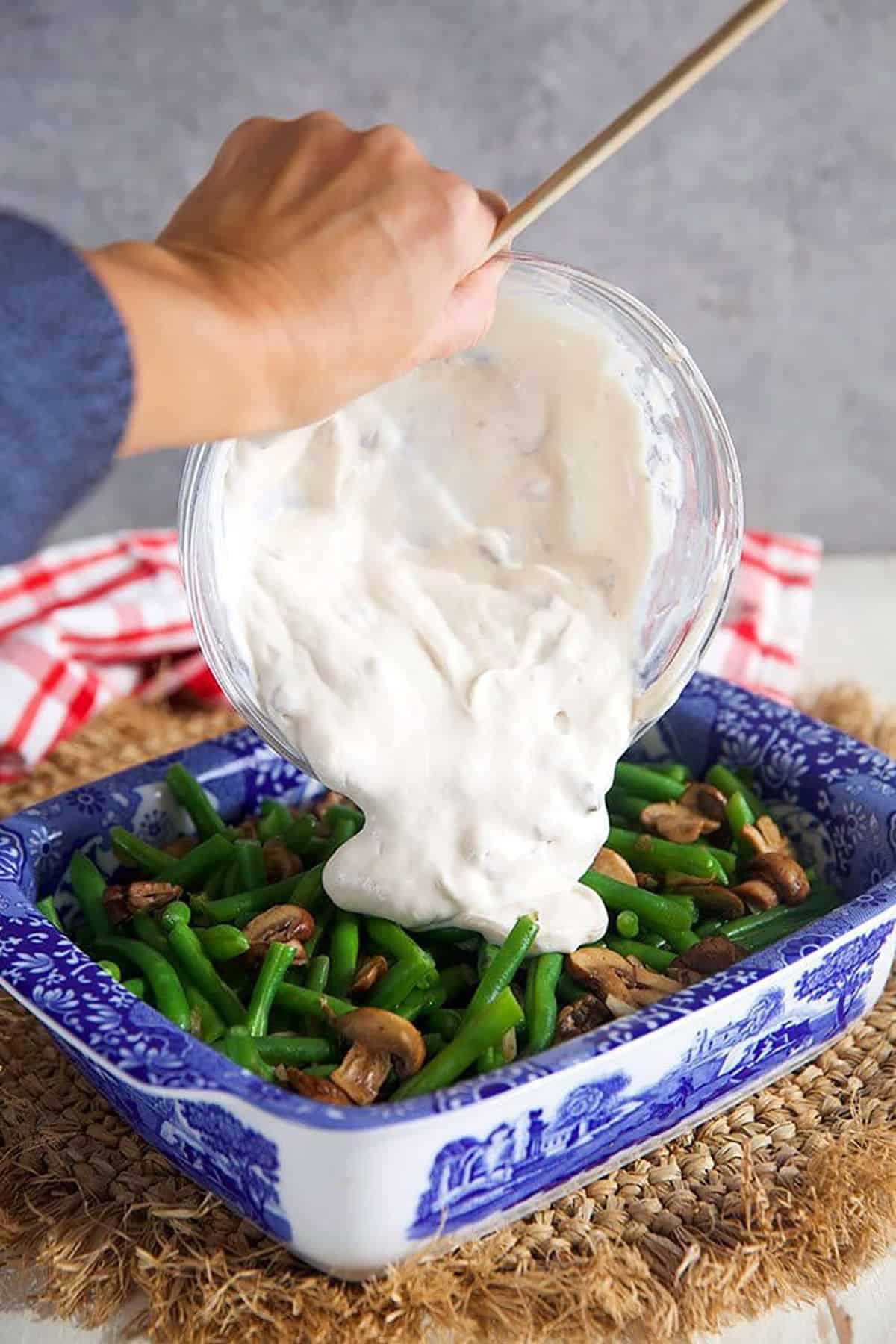 creamy cheese sauce being poured over blanched green beans in a baking dish