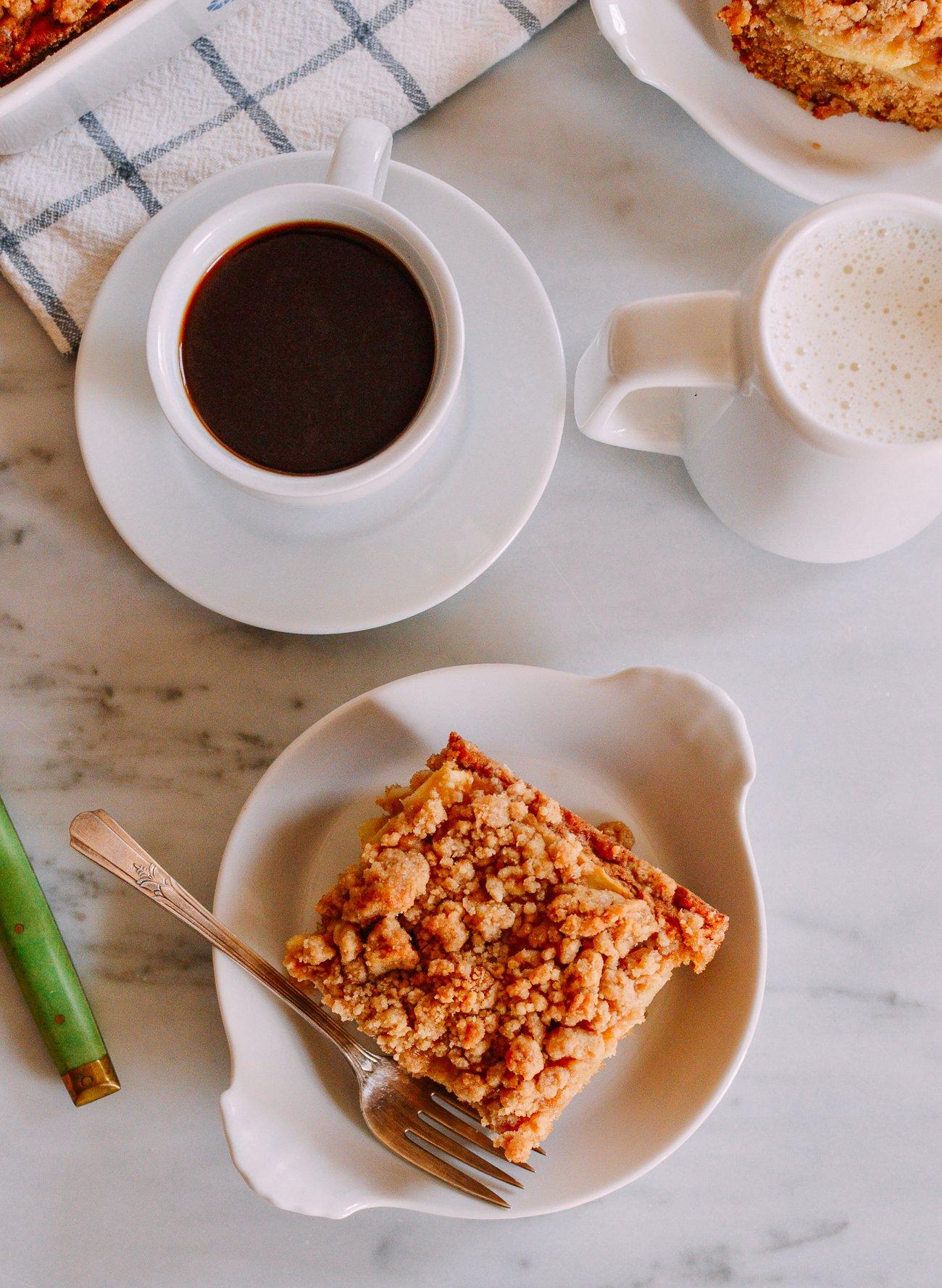 a slice of cinnamon apple cake on a plate, with a fork and a cup of tea next to it