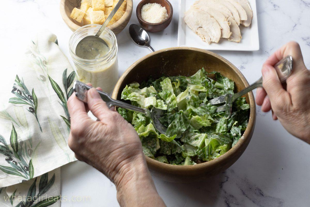 A person tossing a Chicken Caesar Salad in a large bowl.