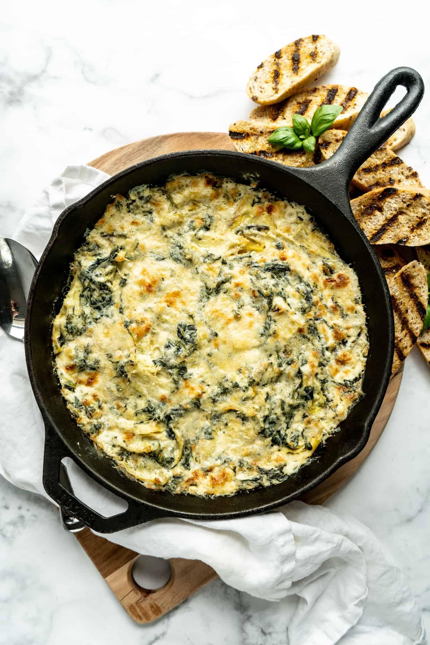 An overhead shot of a baked spinach artichoke dip in a cast iron skillet, surrounded by various dippers like chips, bread, and veggie sticks