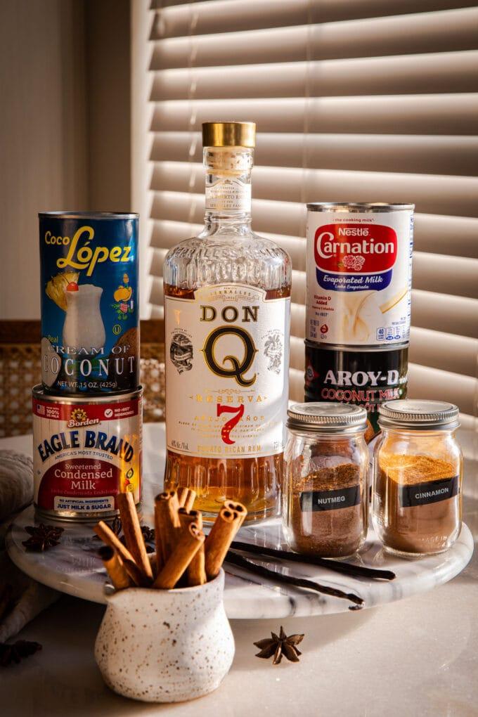 Ingredients for Coquito laid out on a rustic wooden table: cans of milk, coconut cream, rum bottle, spices