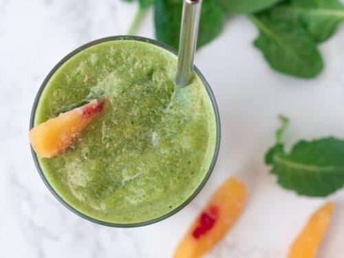 a vibrant green smoothie with peach slices and leafy greens, garnished with mint leaves, in a clear glass on a bright, sunny countertop