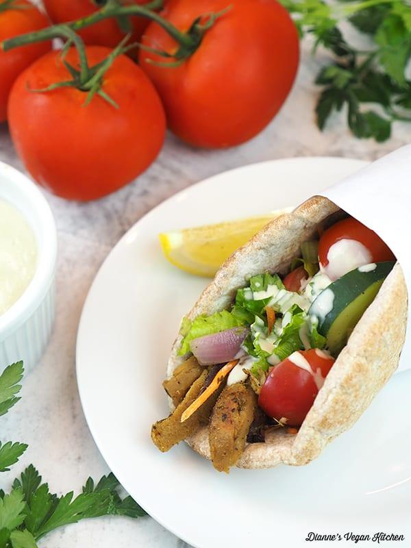 Close-up of a vegan gyro being assembled with seitan, tahini sauce, tomatoes, cucumbers, and red onion