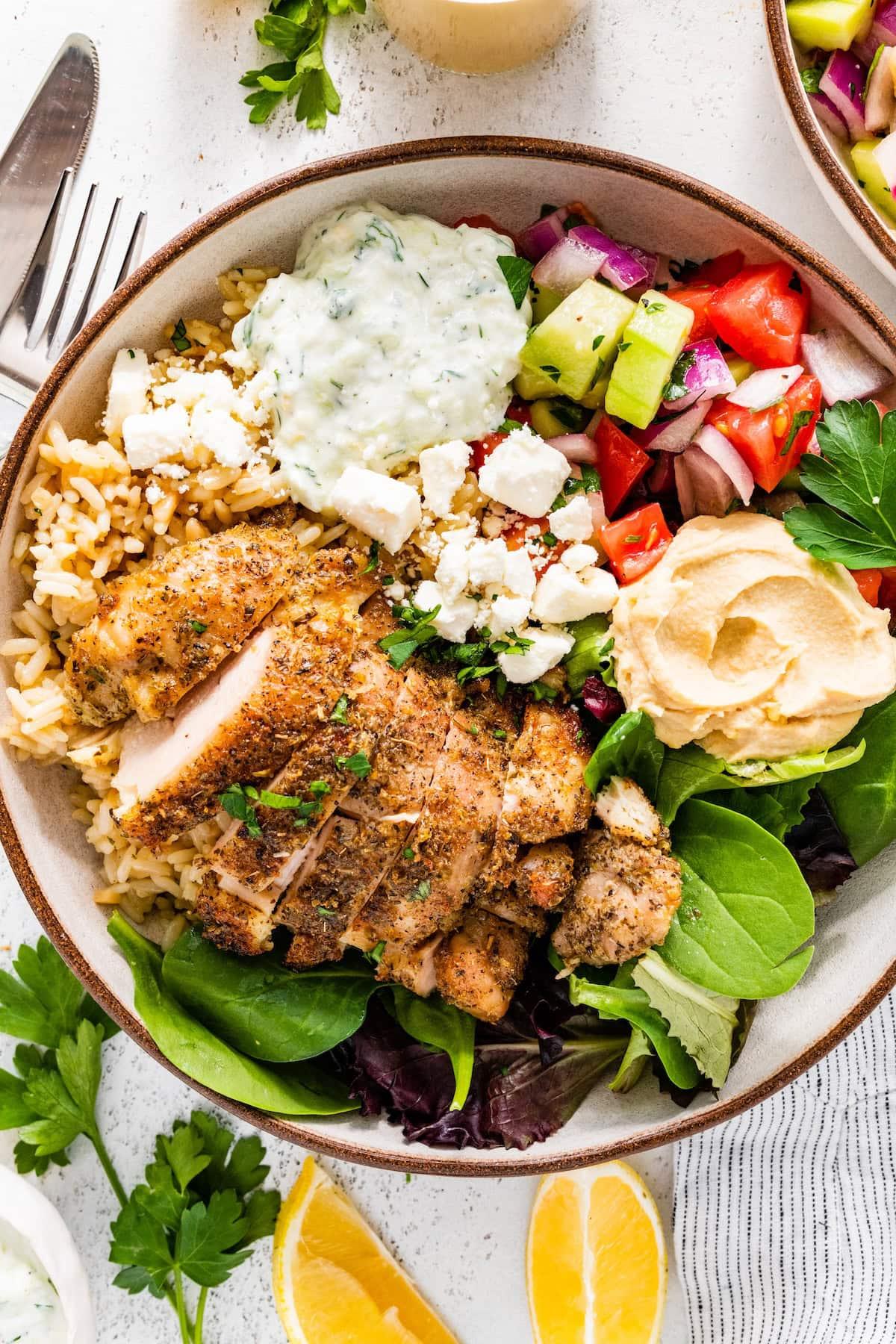 Hands assembling a Greek chicken bowl, adding fresh vegetables and chicken onto a bed of rice