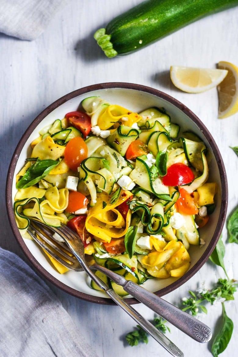 A vibrant bowl of garlic zoodles with zucchini, yellow squash, and feta cheese, garnished with fresh basil.