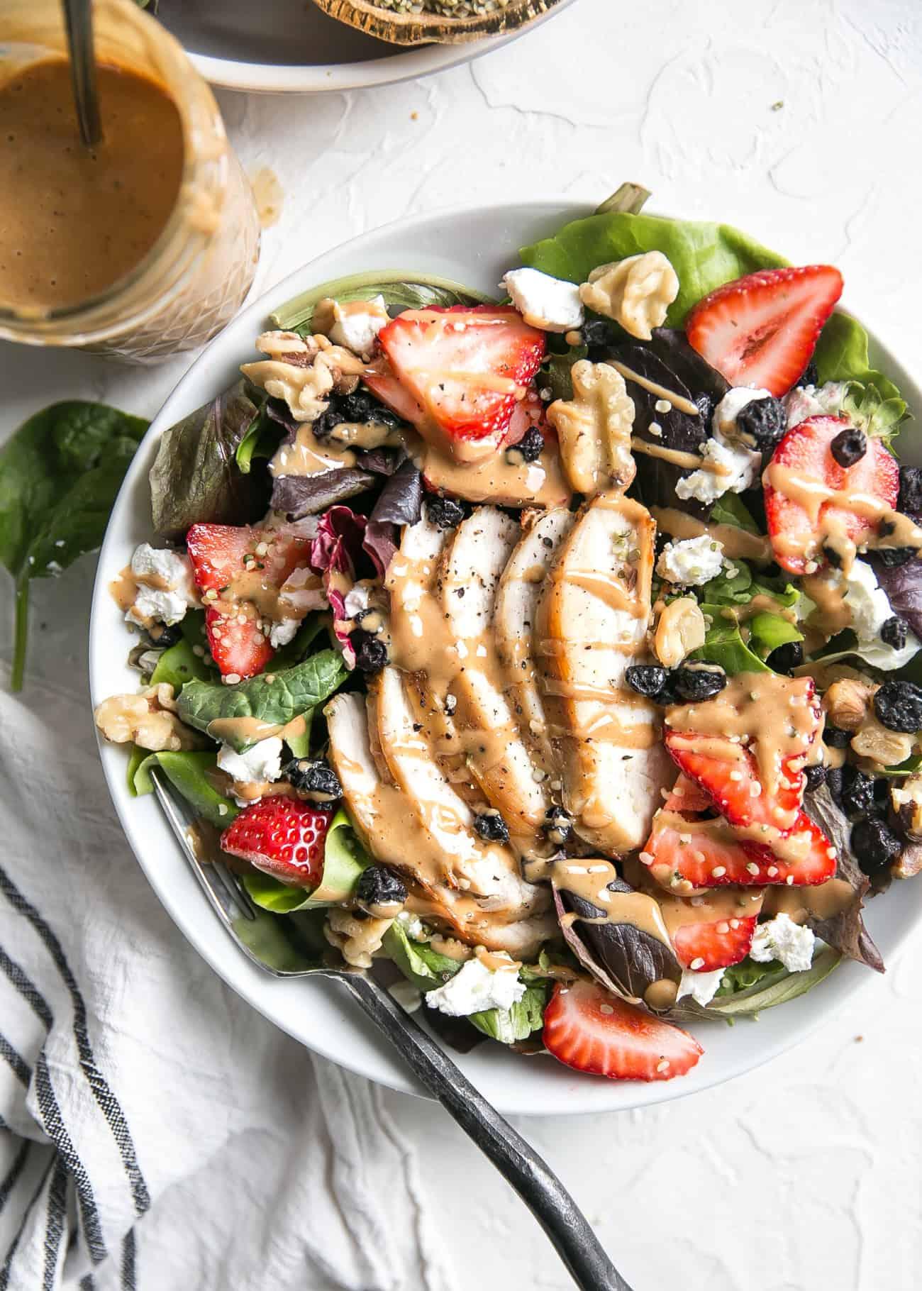 a variety of ingredients being added to a spring strawberry salad: nuts, seeds, goat cheese, grilled chicken