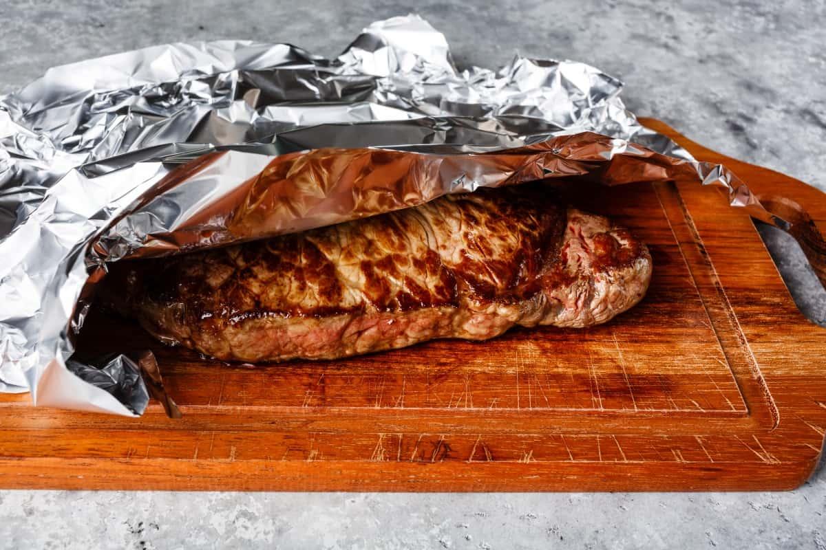 A cooked beef roast resting on a cutting board, lightly tented with foil, with juices pooling slightly.