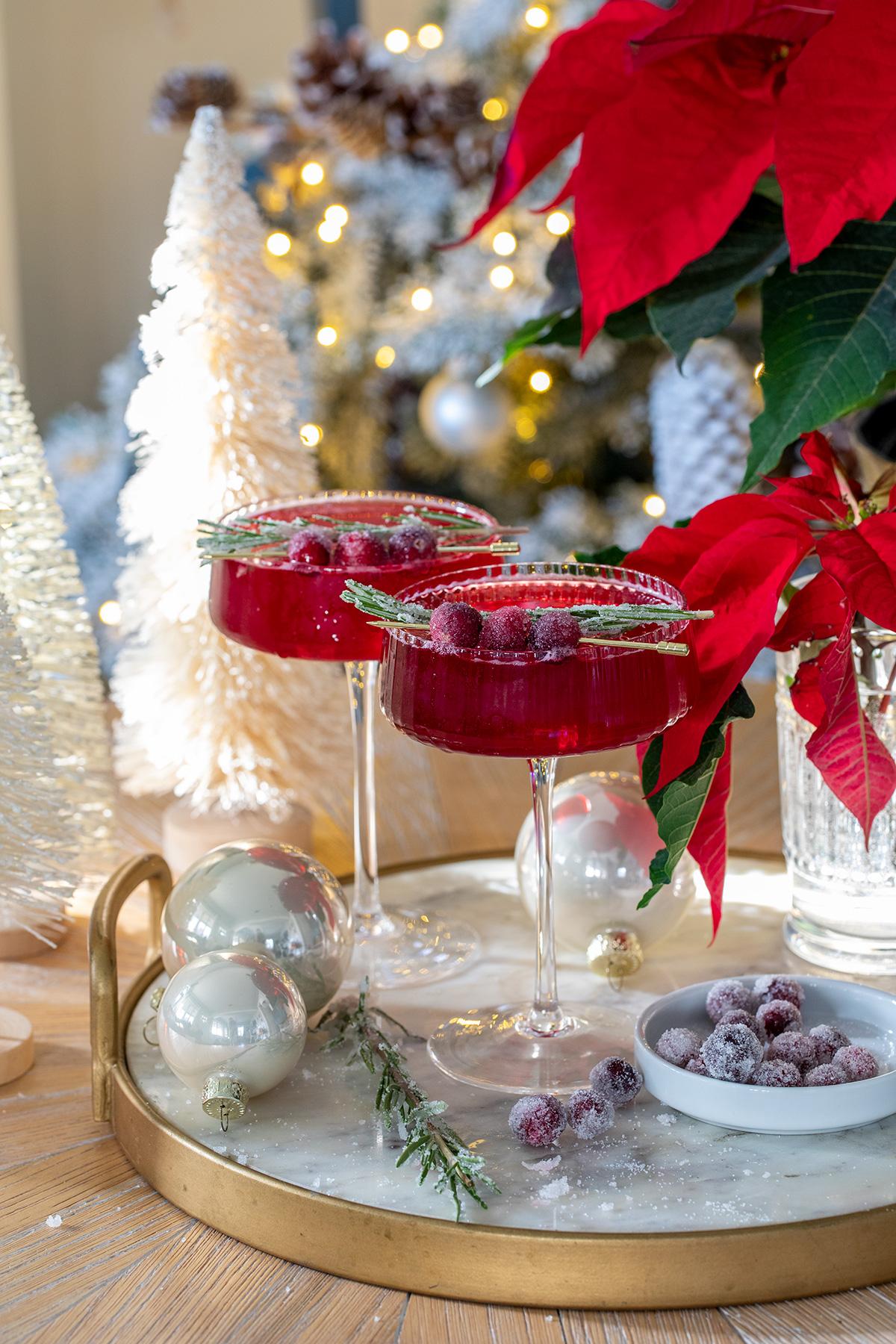 Warm, inviting shot of a holiday table setting with a pitcher of cranberry spritz and several glasses, festive lights in background