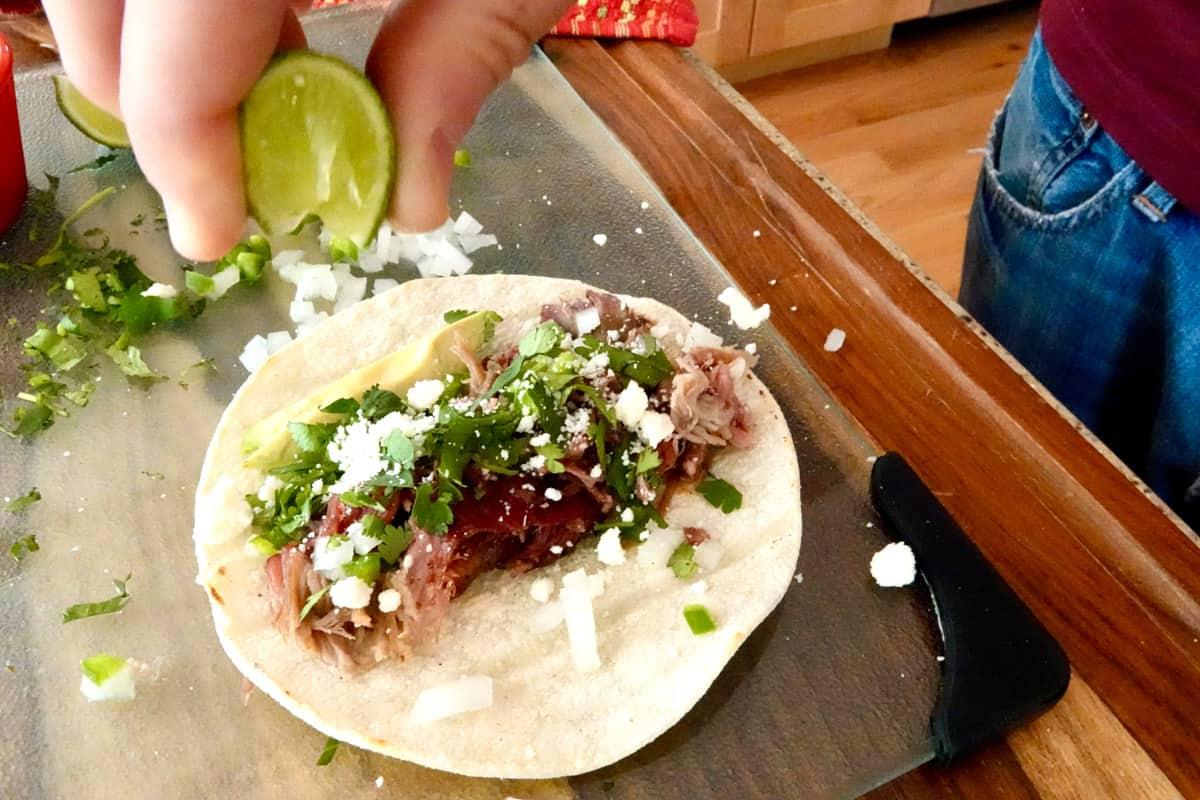 a hand assembling a street taco with pulled pork, onion, and cilantro