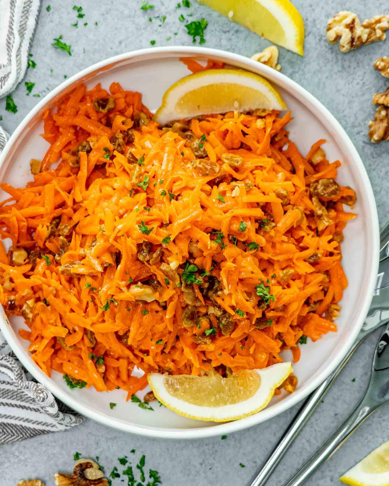 overhead shot of a colorful carrot and raisin salad in a white bowl, surrounded by fresh carrots and ingredients