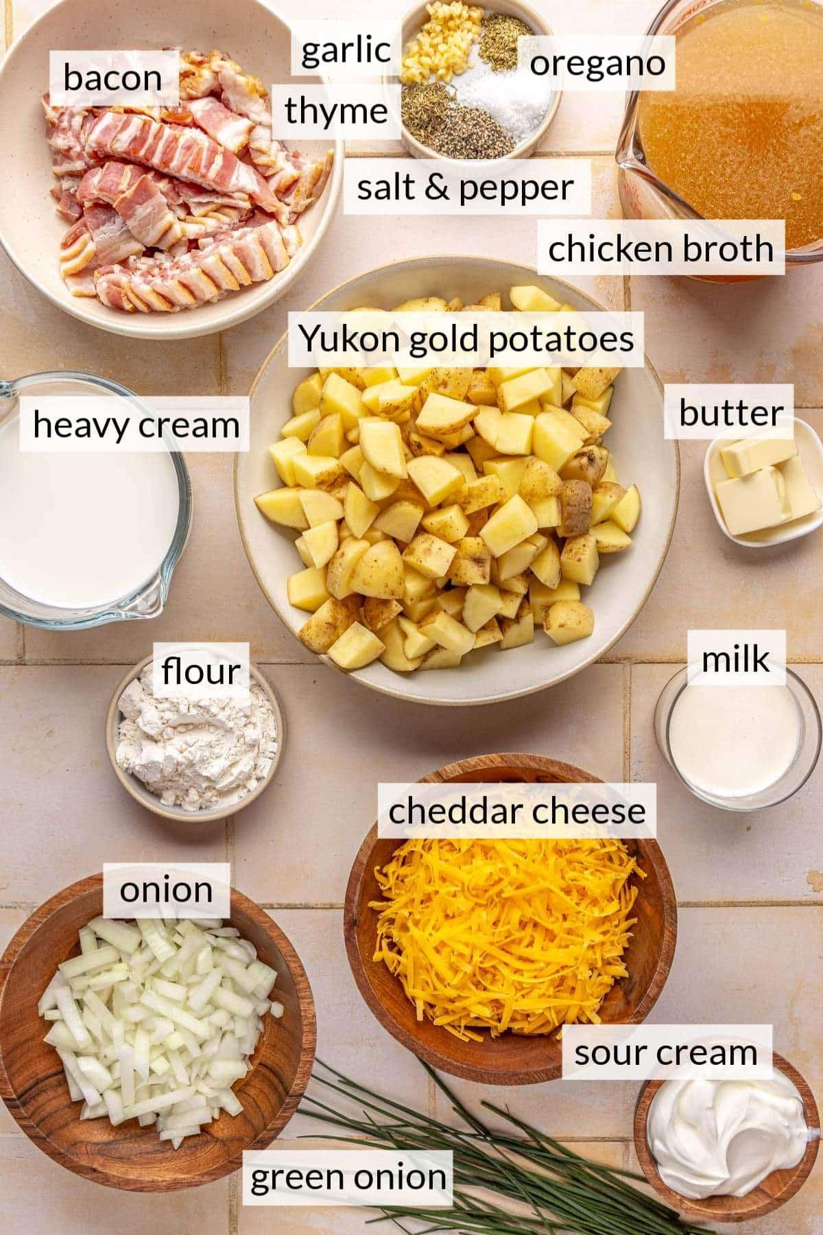 Overhead shot of fresh ingredients for potato soup laid out on a wooden counter: potatoes, cheddar cheese, onion, celery, broth, cream