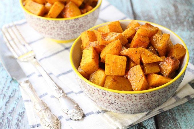 Hands tossing butternut squash cubes with maple syrup and spices in a large mixing bowl