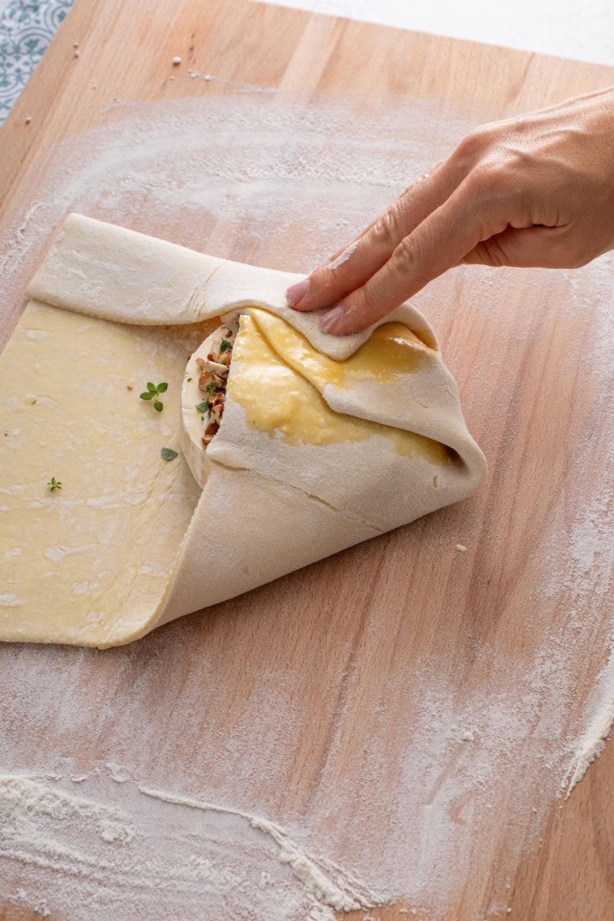 A person gently wrapping a wheel of brie in a sheet of puff pastry on a floured surface