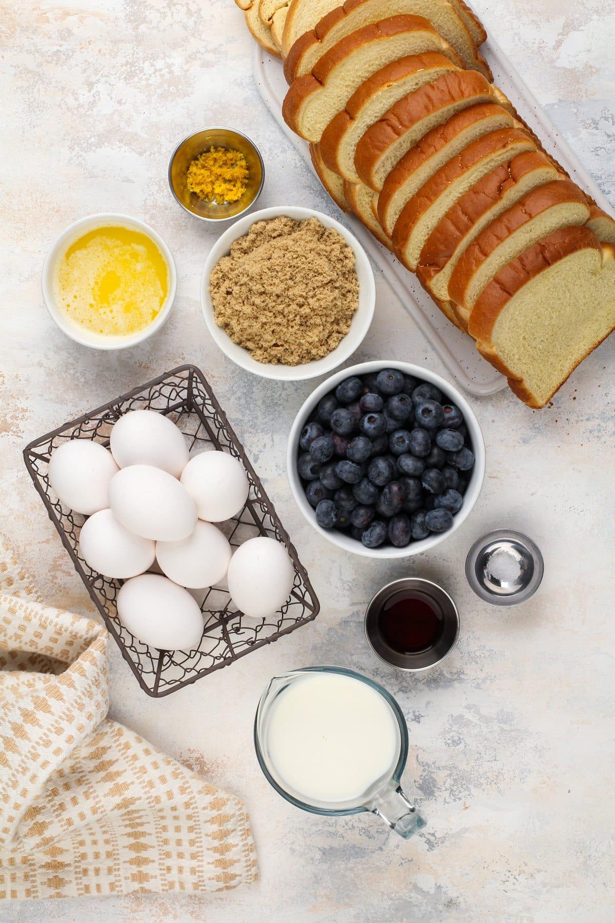 Ingredients for blueberry French toast casserole laid out on a wooden table: bread, eggs, milk, blueberries, streusel ingredients