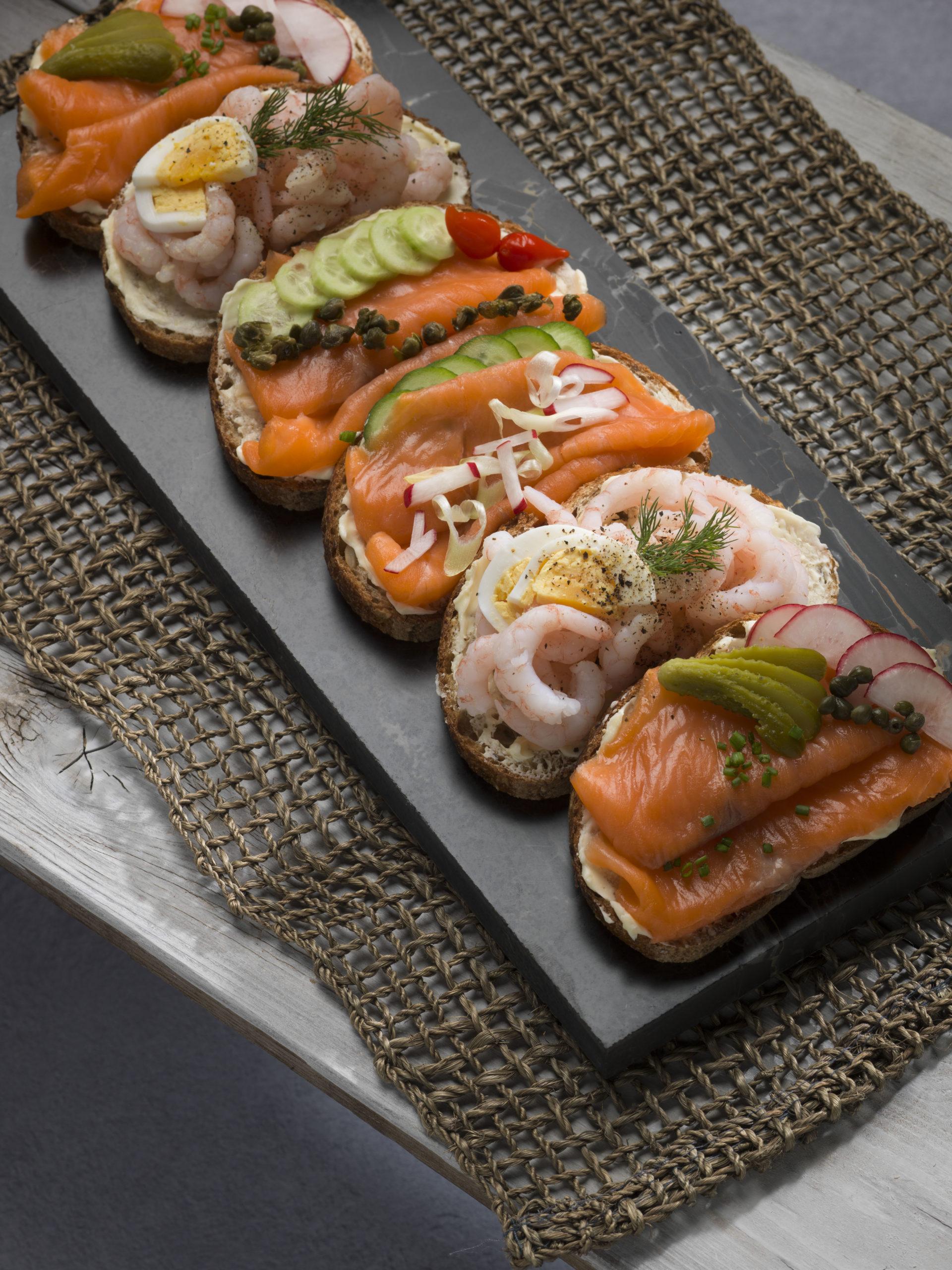 Overhead shot of a beautifully arranged plate of various Smørrebrød, including one prominently featuring smoked eel, with dill sprigs and lemon wedges.
